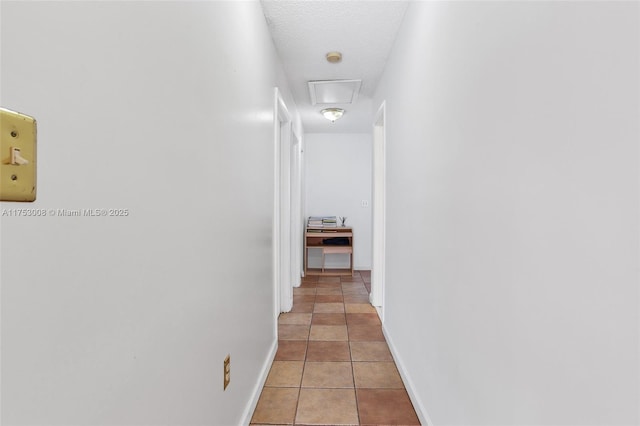 corridor featuring light tile patterned floors, a textured ceiling, attic access, and baseboards