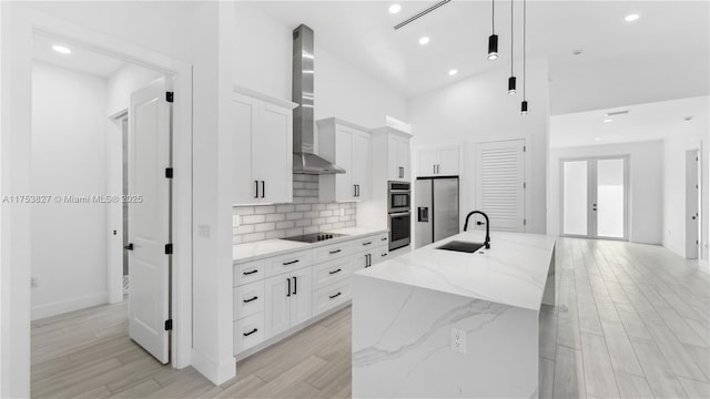 kitchen with french doors, appliances with stainless steel finishes, a sink, wall chimney range hood, and an island with sink