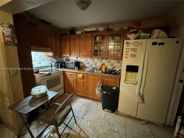 kitchen with light tile patterned floors, under cabinet range hood, white appliances, tasteful backsplash, and brown cabinetry