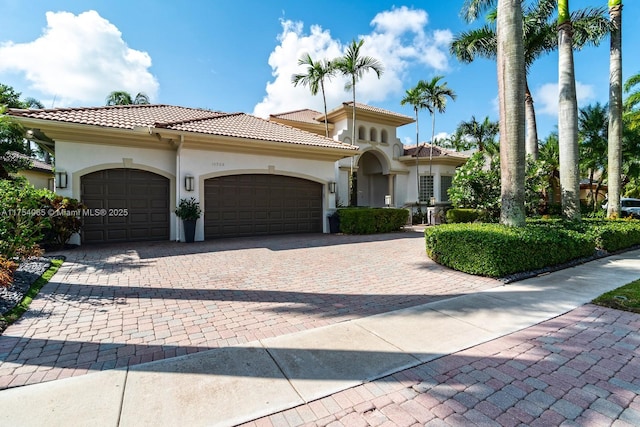 mediterranean / spanish house with stucco siding, an attached garage, a tile roof, and decorative driveway