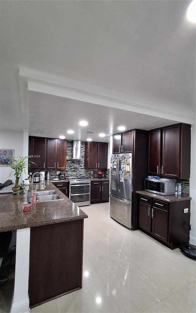 kitchen featuring a peninsula, a sink, appliances with stainless steel finishes, backsplash, and wall chimney exhaust hood