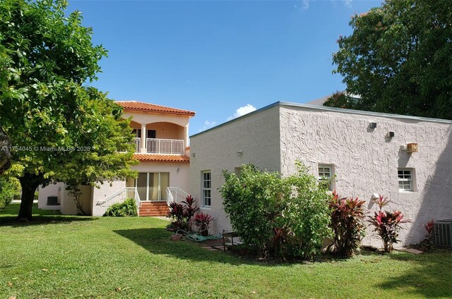 rear view of property featuring a balcony, central air condition unit, a tile roof, a lawn, and stucco siding