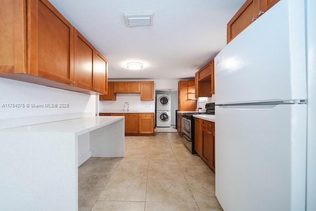 kitchen featuring visible vents, stacked washer / dryer, electric stove, brown cabinets, and freestanding refrigerator