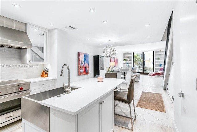 kitchen featuring open floor plan, wall chimney range hood, premium stove, tasteful backsplash, and an island with sink