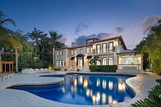 back of house at dusk with an outdoor pool, a patio, a balcony, a chimney, and an outdoor hangout area