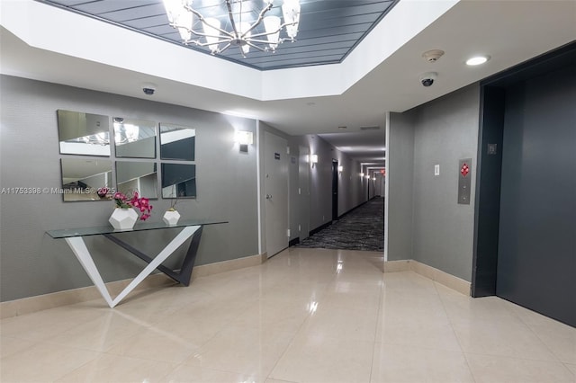 hall featuring a notable chandelier, a raised ceiling, elevator, tile patterned flooring, and baseboards