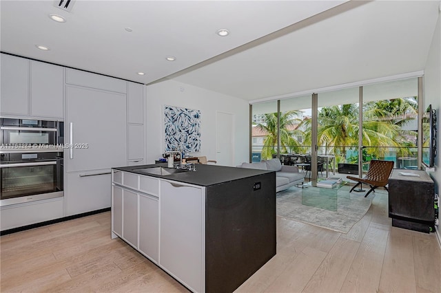 kitchen with white cabinets, dark countertops, expansive windows, light wood-type flooring, and a sink