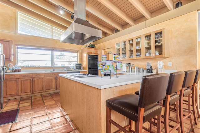 kitchen featuring a breakfast bar area, light countertops, dobule oven black, glass insert cabinets, and exhaust hood