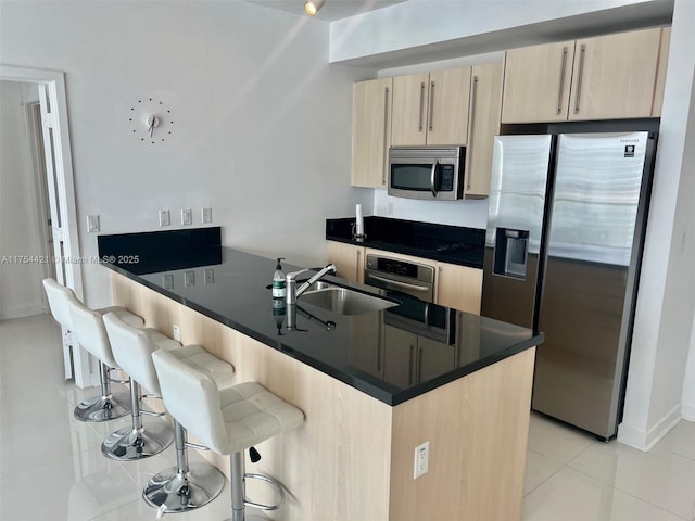 kitchen featuring a kitchen bar, light brown cabinets, a sink, dark countertops, and stainless steel appliances