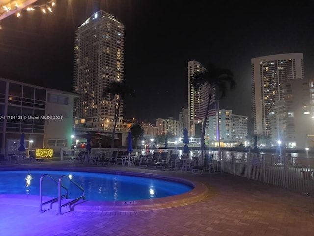 pool at twilight featuring a view of city lights, fence, and a community pool