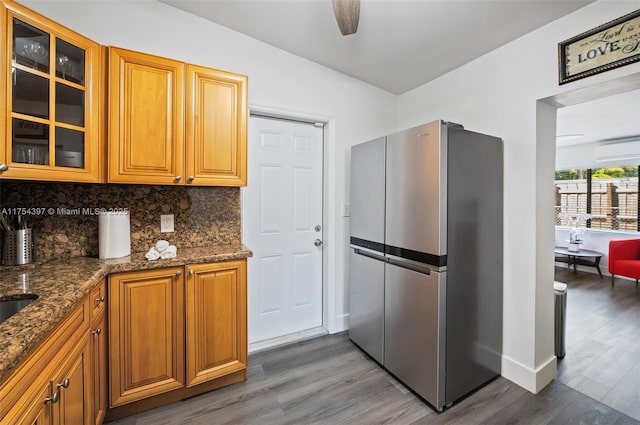 kitchen with tasteful backsplash, glass insert cabinets, freestanding refrigerator, wood finished floors, and dark stone counters