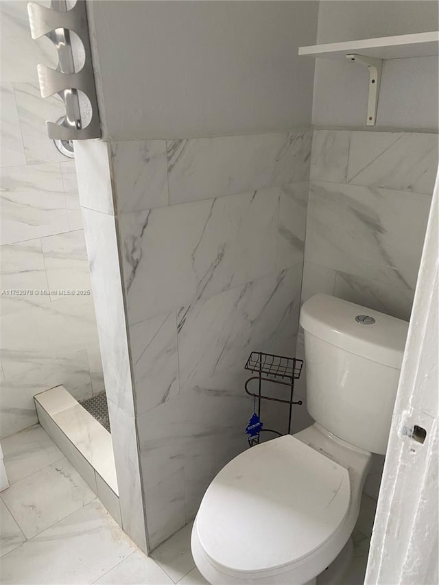 bathroom with toilet, marble finish floor, a tile shower, and tile walls