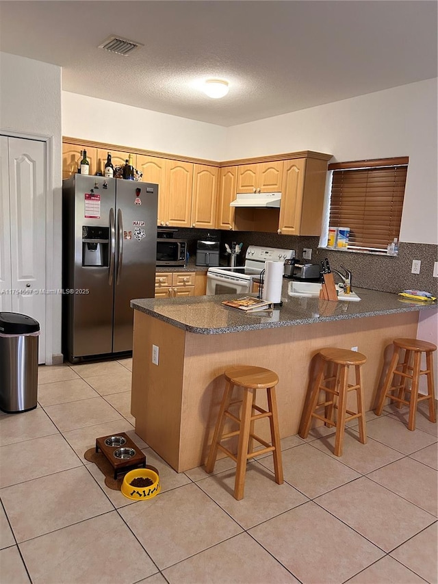 kitchen featuring tasteful backsplash, appliances with stainless steel finishes, a peninsula, light brown cabinets, and light tile patterned flooring