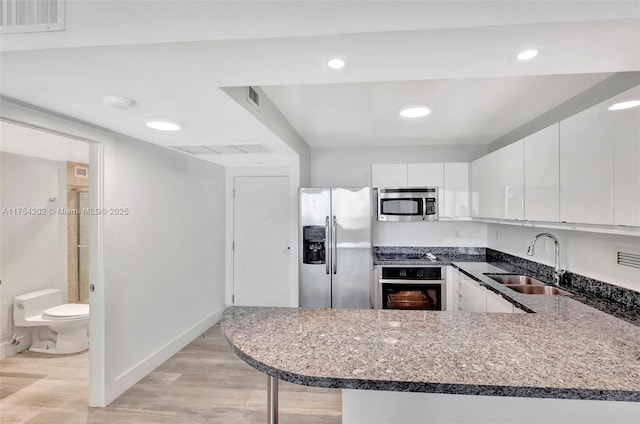 kitchen with stainless steel appliances, a peninsula, a sink, light wood-type flooring, and dark stone countertops