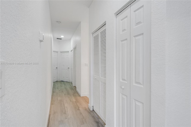 corridor with light wood-style flooring, visible vents, baseboards, and a textured wall