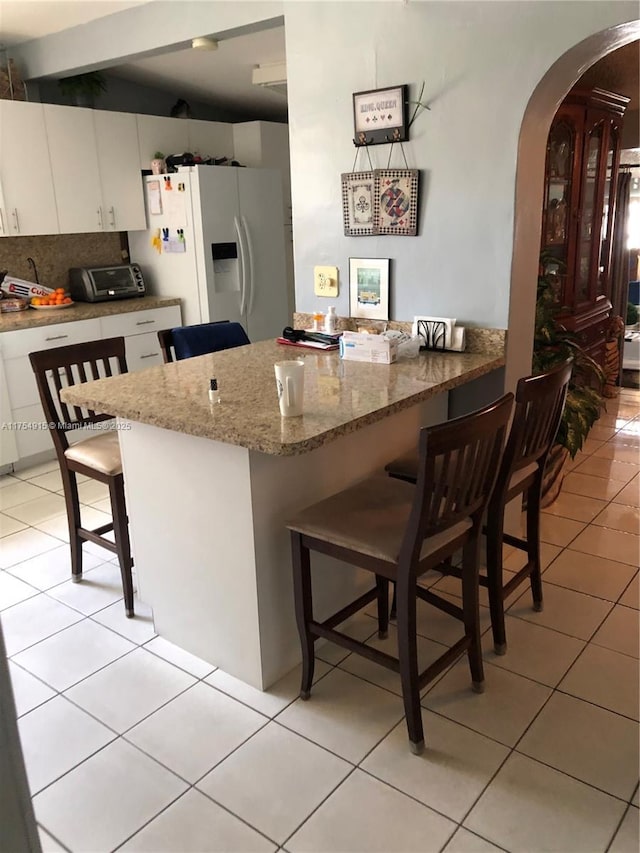 kitchen featuring arched walkways, light tile patterned floors, white cabinets, white fridge with ice dispenser, and a kitchen bar