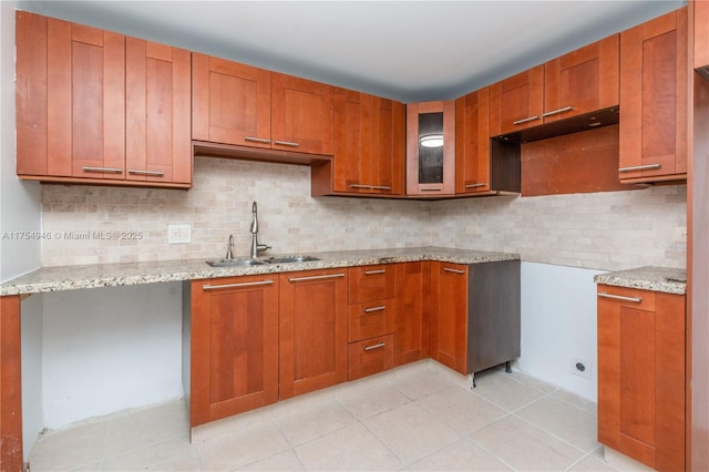 kitchen featuring light stone counters, brown cabinets, tasteful backsplash, glass insert cabinets, and a sink