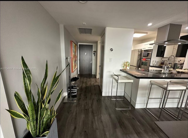 kitchen featuring visible vents, dark wood finished floors, wall chimney range hood, and a kitchen breakfast bar