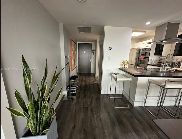 kitchen featuring wall chimney range hood, dark wood-style flooring, visible vents, and a kitchen breakfast bar