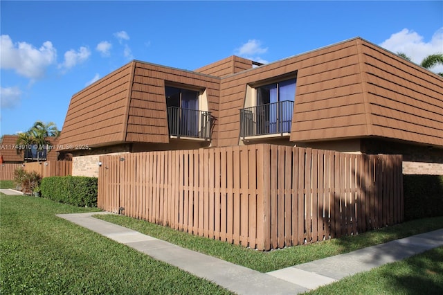 view of side of home with mansard roof, a balcony, brick siding, fence, and a yard