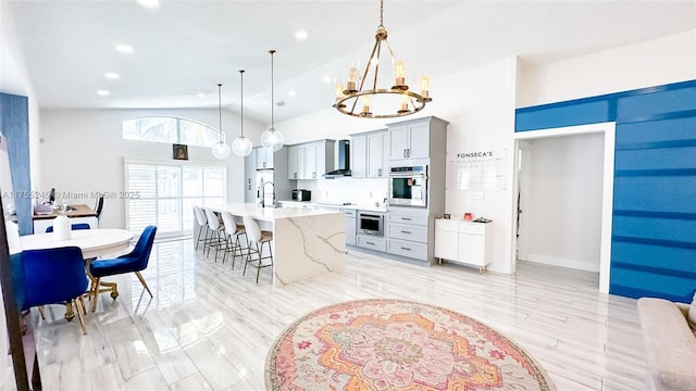 kitchen with an island with sink, wall chimney exhaust hood, oven, a kitchen bar, and a chandelier