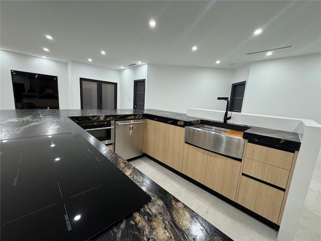 kitchen featuring light brown cabinetry, dark stone countertops, recessed lighting, stainless steel appliances, and a sink