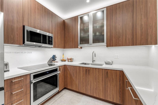 kitchen featuring stainless steel appliances, a sink, brown cabinetry, modern cabinets, and glass insert cabinets