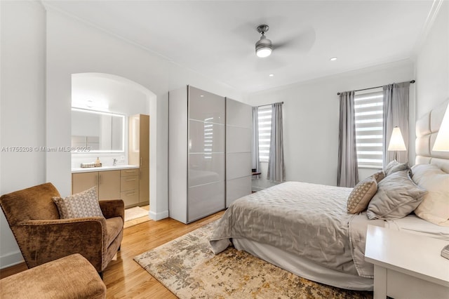 bedroom with arched walkways, a ceiling fan, ornamental molding, ensuite bathroom, and light wood-type flooring