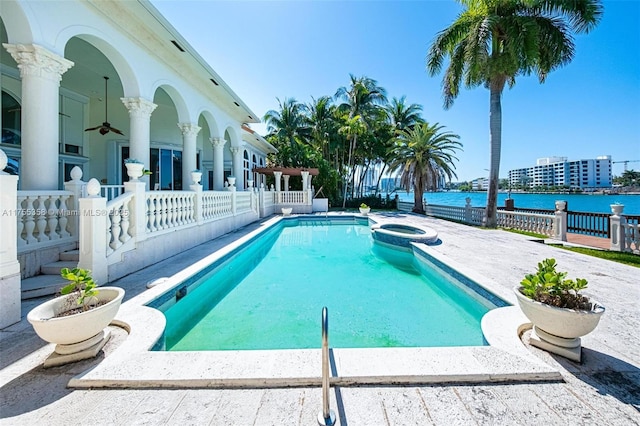 view of swimming pool featuring a ceiling fan, fence, a pool with connected hot tub, a water view, and a patio area