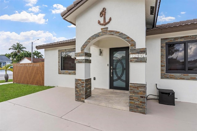 view of exterior entry with stone siding, a tile roof, fence, and stucco siding