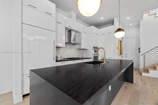 kitchen with modern cabinets, a sink, dark countertops, wall chimney exhaust hood, and light wood finished floors