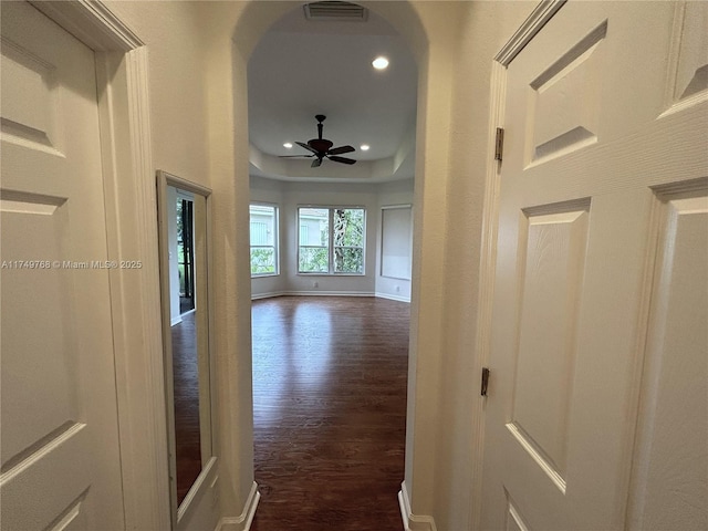 corridor featuring arched walkways, dark wood-type flooring, visible vents, baseboards, and a tray ceiling