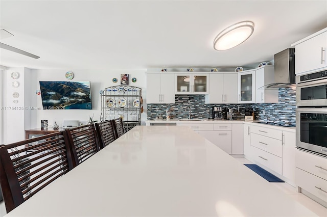 kitchen featuring tasteful backsplash, wall chimney exhaust hood, light countertops, stainless steel double oven, and a sink