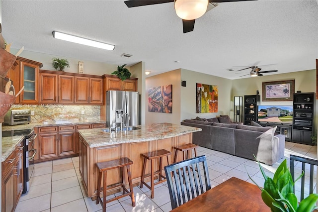 kitchen featuring stainless steel appliances, backsplash, brown cabinetry, a kitchen island with sink, and a kitchen breakfast bar