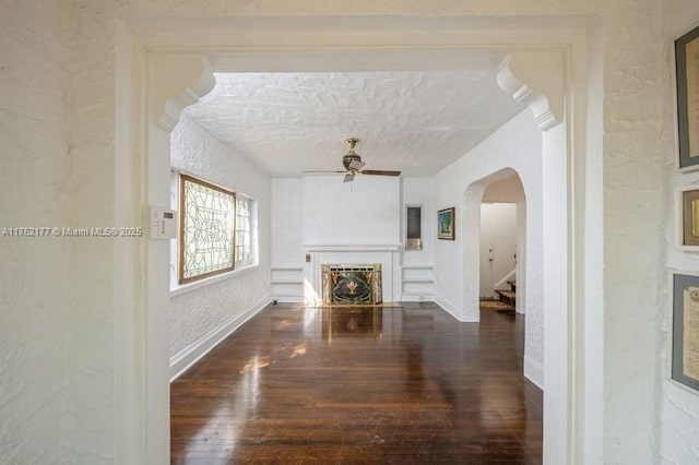 unfurnished living room with arched walkways, a textured wall, wood finished floors, a fireplace with flush hearth, and a ceiling fan