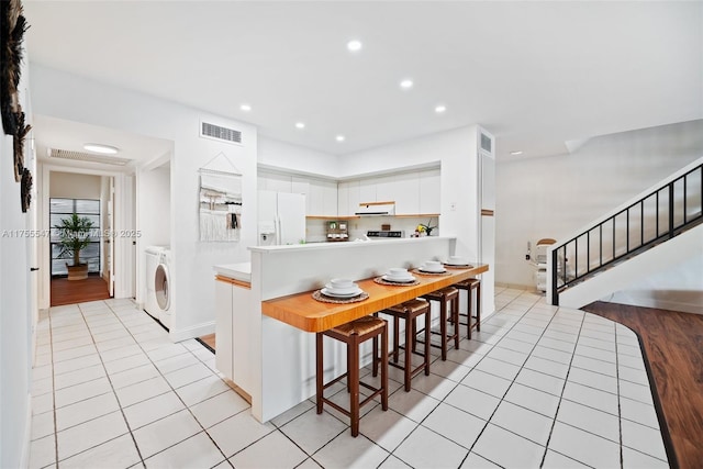 kitchen with light tile patterned flooring, under cabinet range hood, white cabinets, white fridge with ice dispenser, and washer / dryer