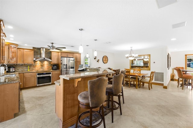 kitchen featuring wall chimney exhaust hood, appliances with stainless steel finishes, brown cabinets, a kitchen bar, and a sink