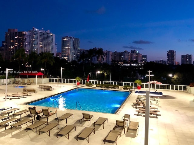 pool at night with a view of city lights, a patio, and a community pool