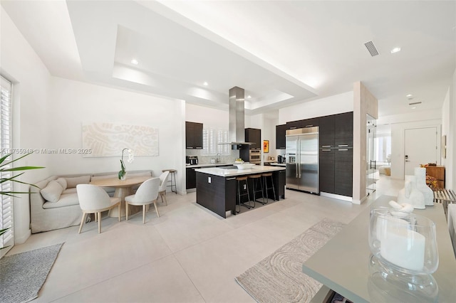 kitchen with stainless steel built in fridge, light countertops, wall chimney range hood, a center island, and a raised ceiling