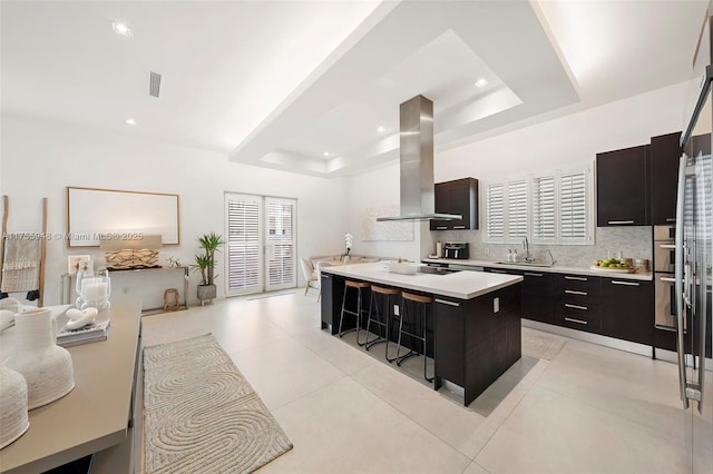 kitchen with a kitchen island, light countertops, ventilation hood, tasteful backsplash, and a raised ceiling