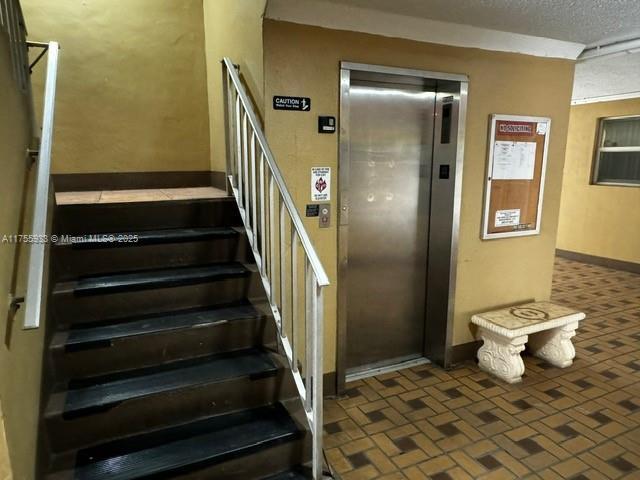 staircase with a textured ceiling, brick floor, baseboards, and elevator