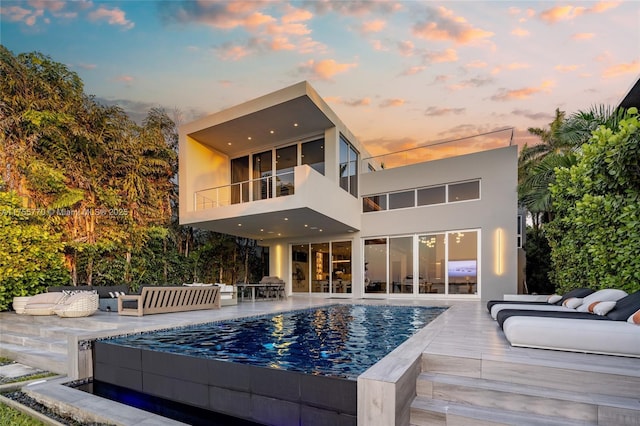 back of property at dusk with a patio, a balcony, an outdoor living space, an outdoor pool, and stucco siding