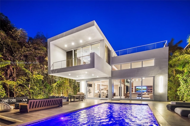 back of house at twilight with an outdoor pool, a patio area, a balcony, outdoor lounge area, and stucco siding