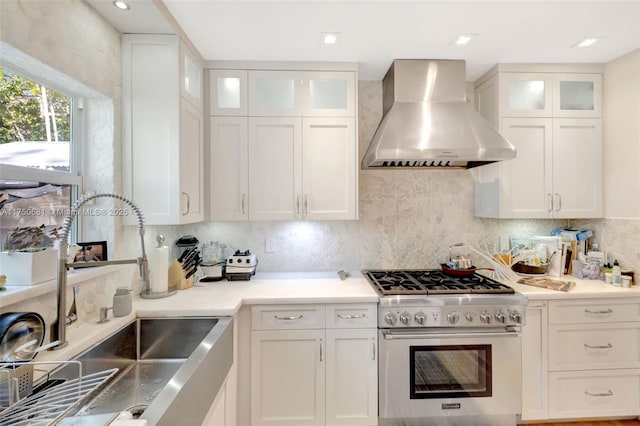 kitchen with stainless steel stove, white cabinetry, a sink, and wall chimney range hood