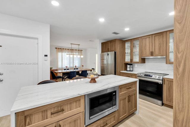 kitchen with appliances with stainless steel finishes, glass insert cabinets, light wood-style flooring, and light stone countertops