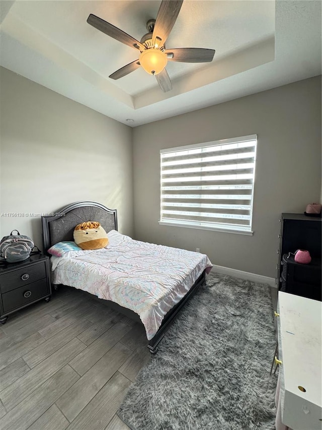 bedroom featuring light wood-type flooring, a tray ceiling, baseboards, and a ceiling fan