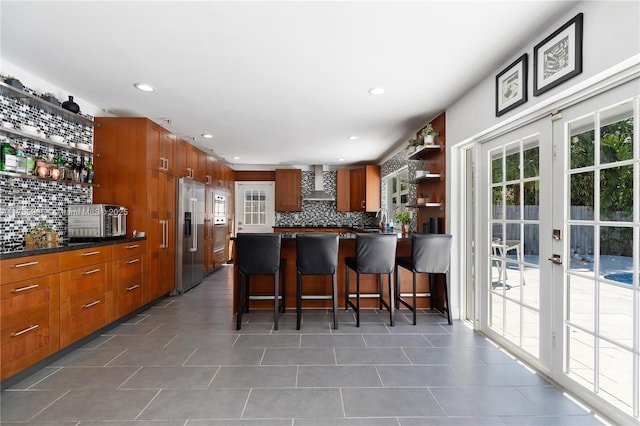 kitchen with open shelves, wall chimney range hood, brown cabinetry, and high quality fridge