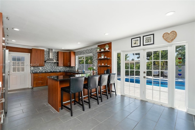 kitchen featuring a kitchen breakfast bar, decorative backsplash, wall chimney exhaust hood, brown cabinetry, and dark countertops