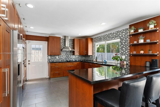 kitchen with decorative backsplash, brown cabinetry, a peninsula, wall chimney range hood, and a sink