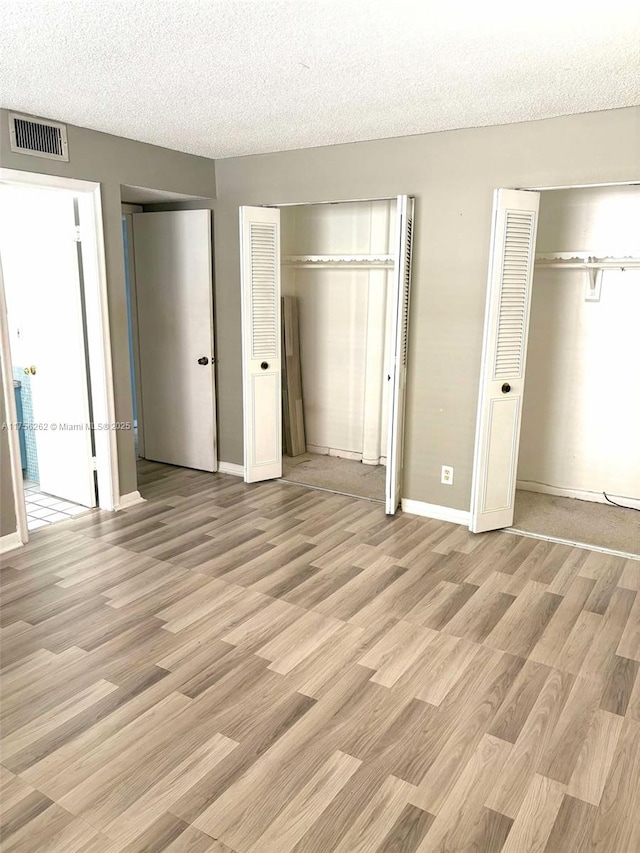 unfurnished bedroom with baseboards, visible vents, a textured ceiling, light wood-type flooring, and two closets